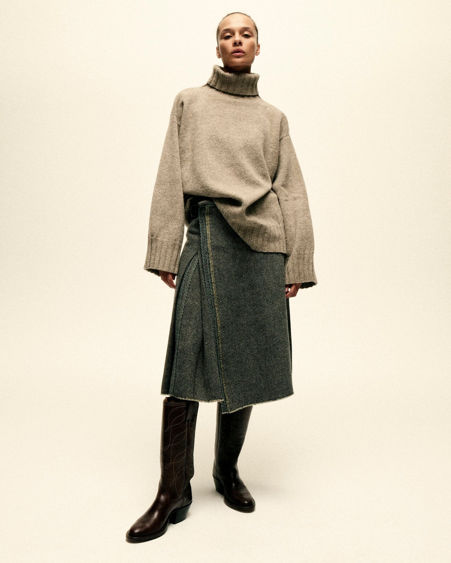 A model stands in front of a white backdrop wearing a turtleneck sweater, kilt, and cowboy boots.