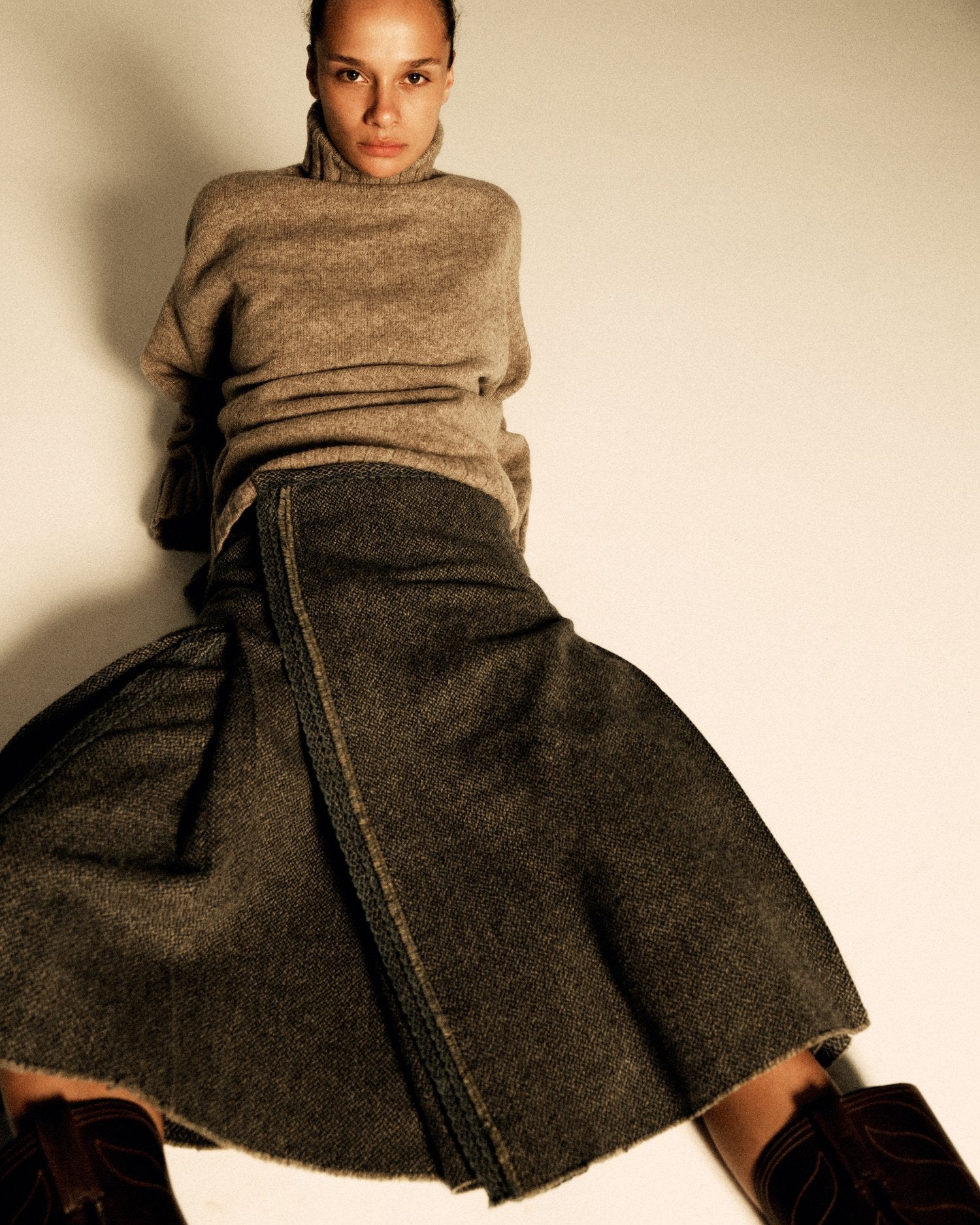 A model lays on the floor wearing a turtleneck sweater, a kilt, and cowboy boots.