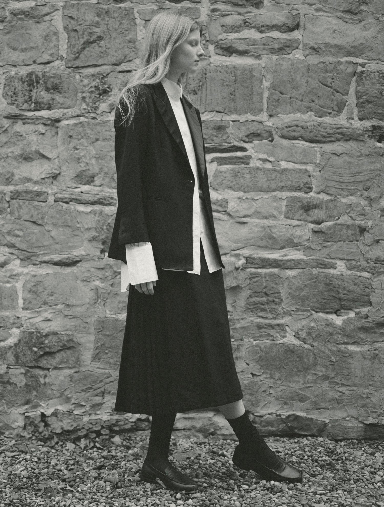 Black and white image of a model standing in front of a stone wall wearing a kilt and matching tuxedo jacket