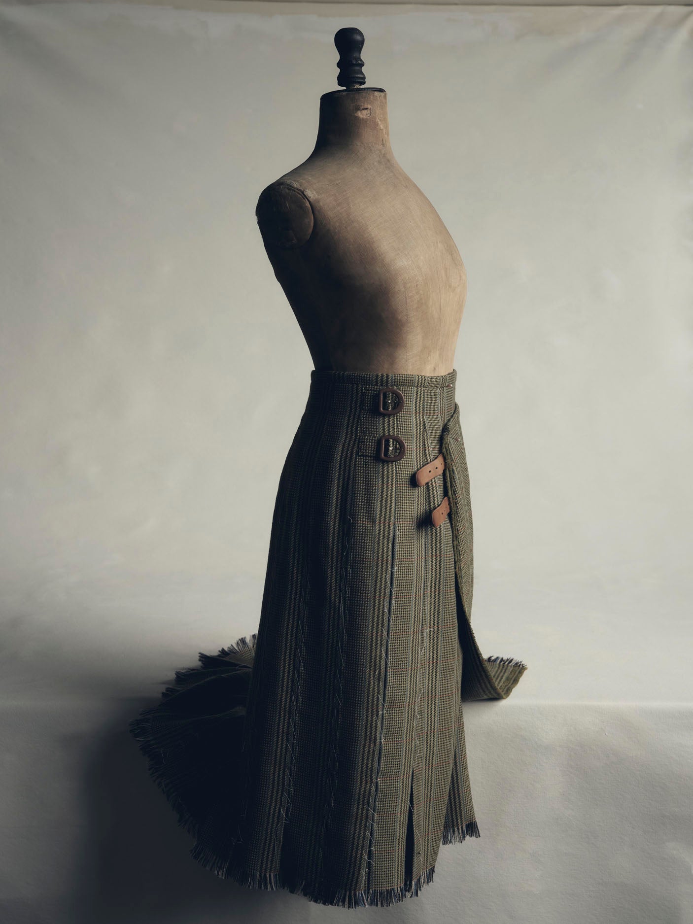 An antique mannequin is dressed in a striped kilt with leather strap detailing in front of a white backdrop.