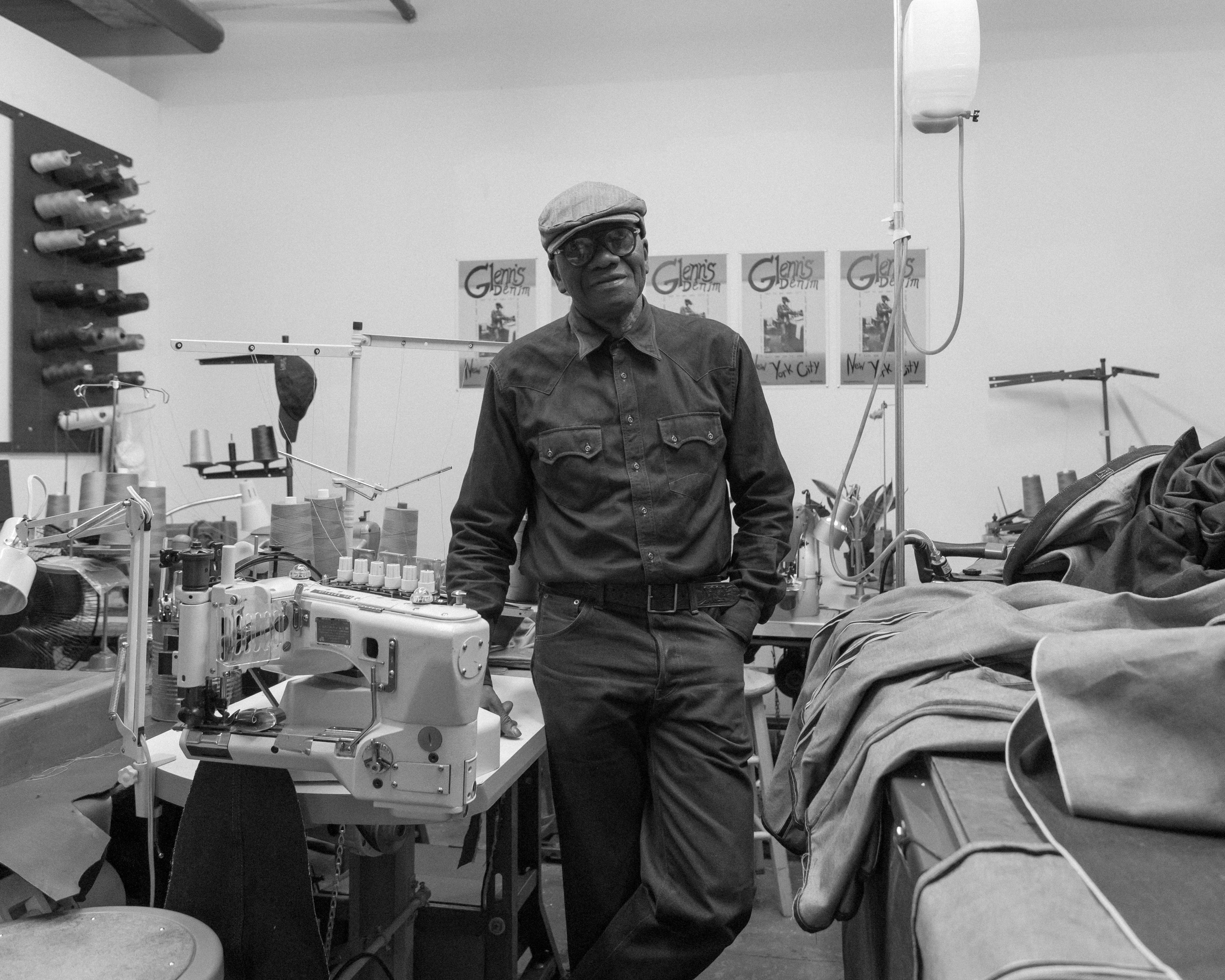 A man poses with hand in pocket standing in the middle of his work room surrounded by sewing machines and fabric.