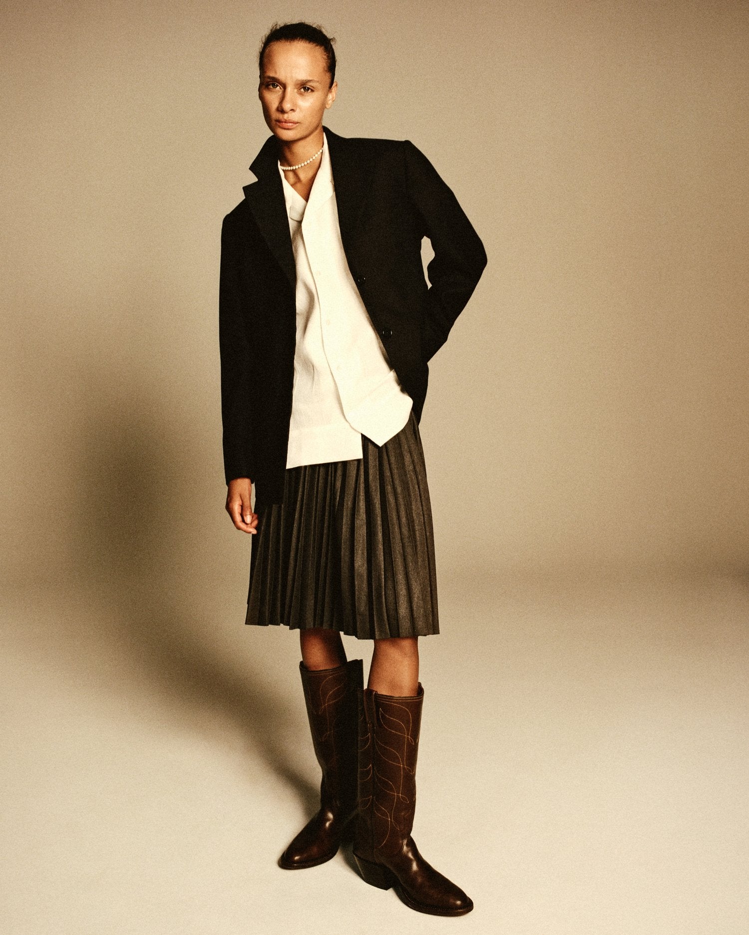 A model wearing a blue blazer, skirt, and cowboy boots stands posing in front of a cream colored background.