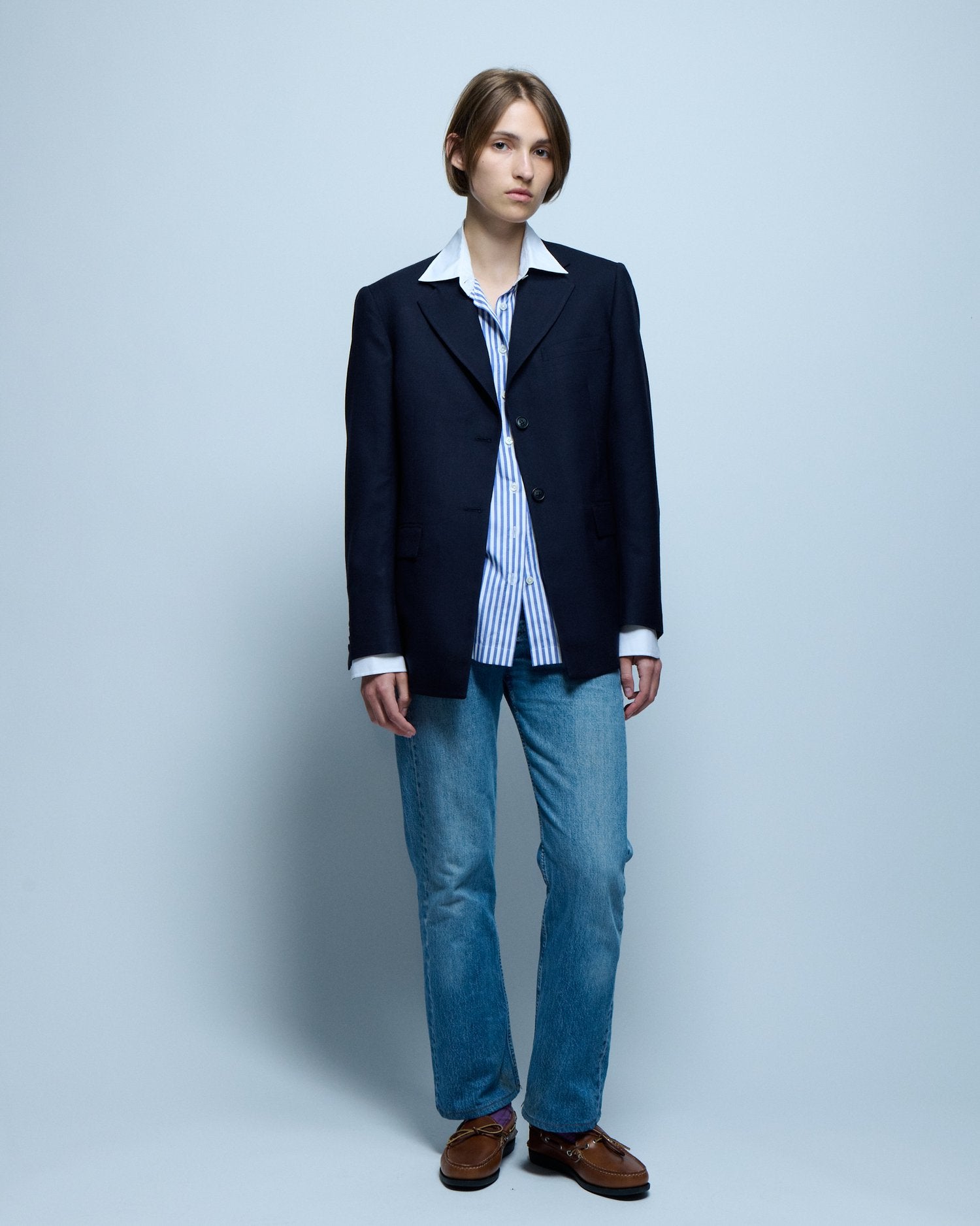 A model wearing a blue blazer, jeans, and boat shoes stands posing in front of a white background.