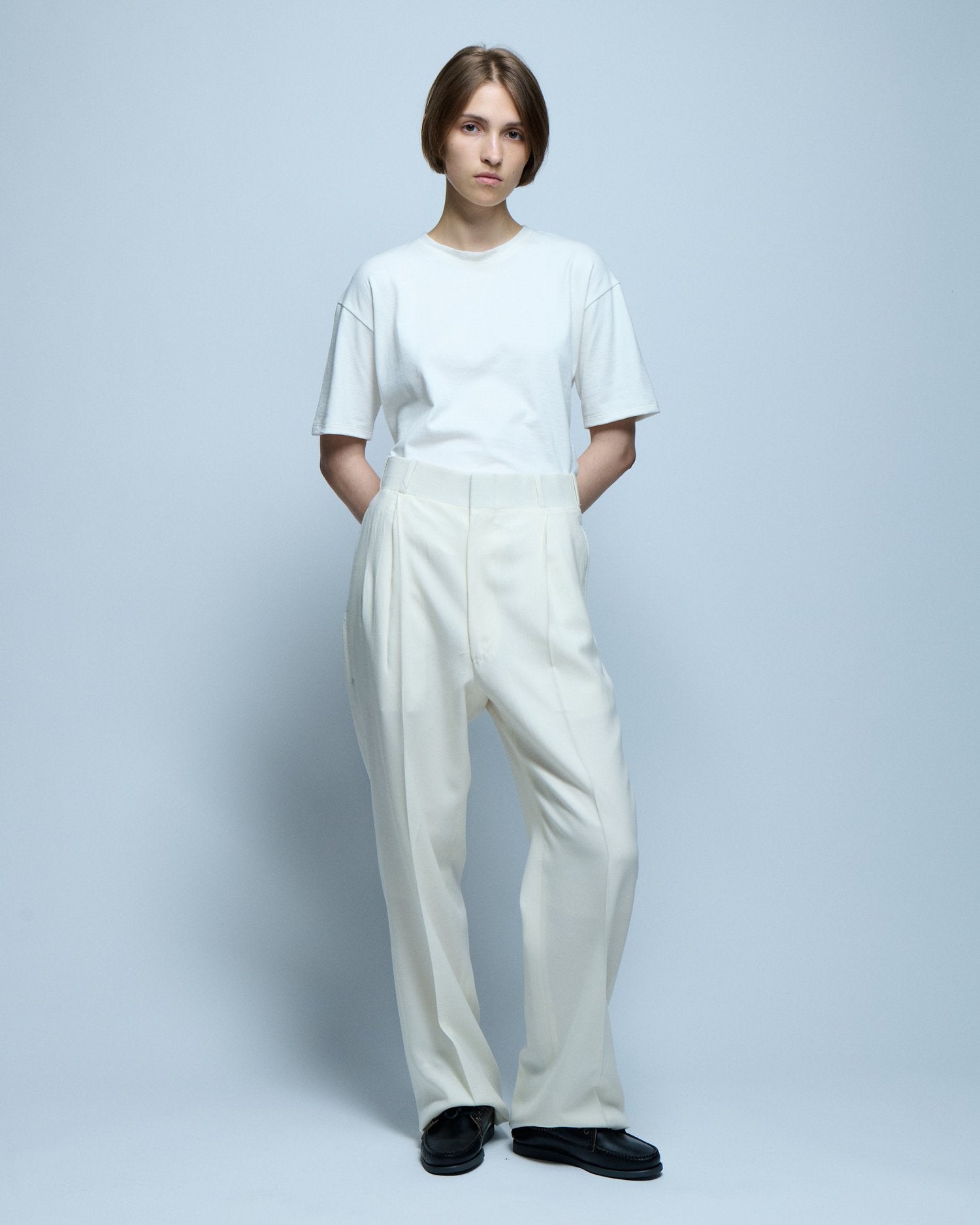 A model stands posing with her hands behind her back wearing a white t-shirt and white pleated trousers.