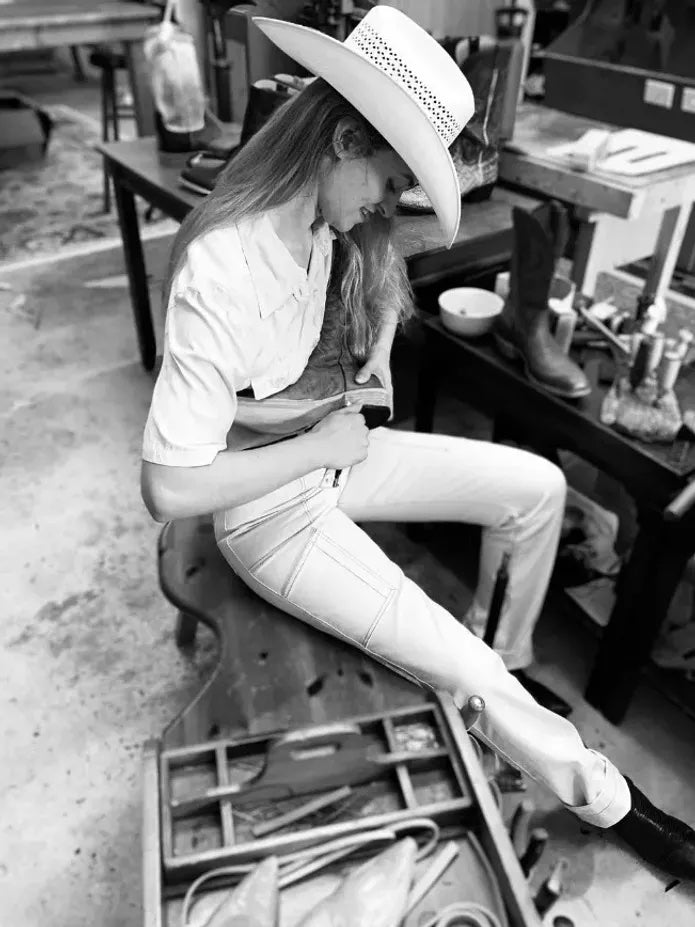 A black and white action shot of Holly Henry sitting on a work bench wearing a cowboy hat while she crafts a handmade boot using a leather making tool.