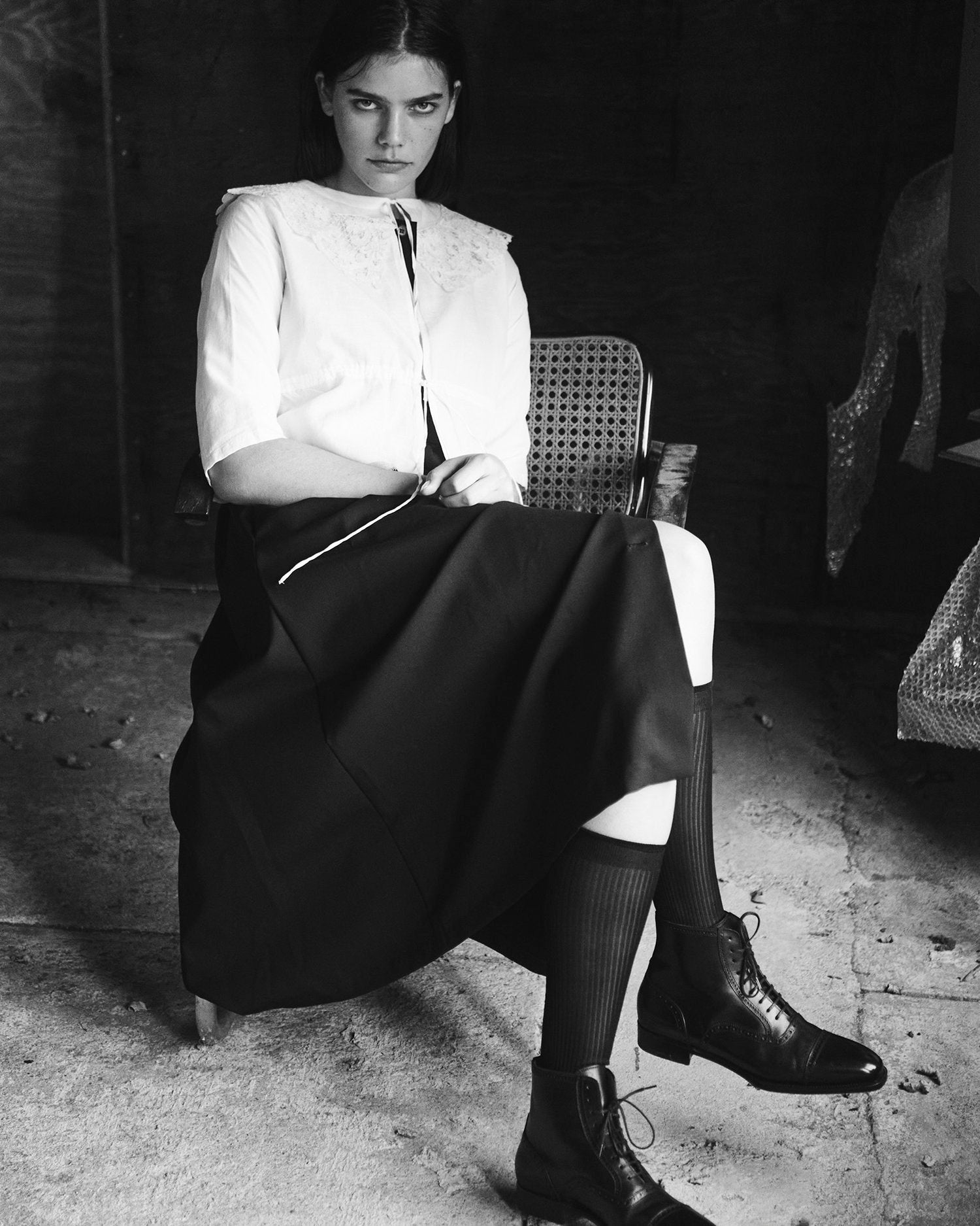 Black and white image of a model sitting cross legged, wearing a white chemise top, black skirt, and black boots.