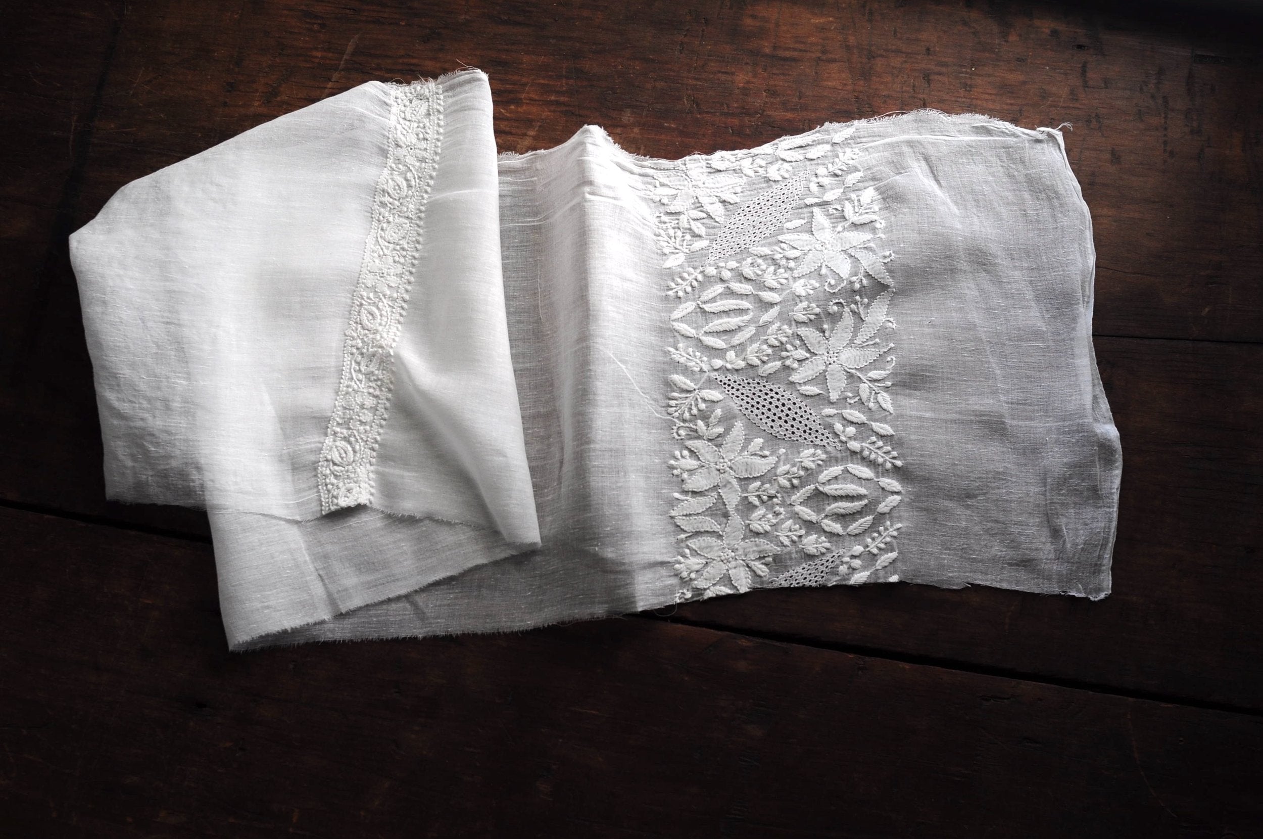 A delicate piece of white embroidered fabric lays partially folded on top of a wooden table.