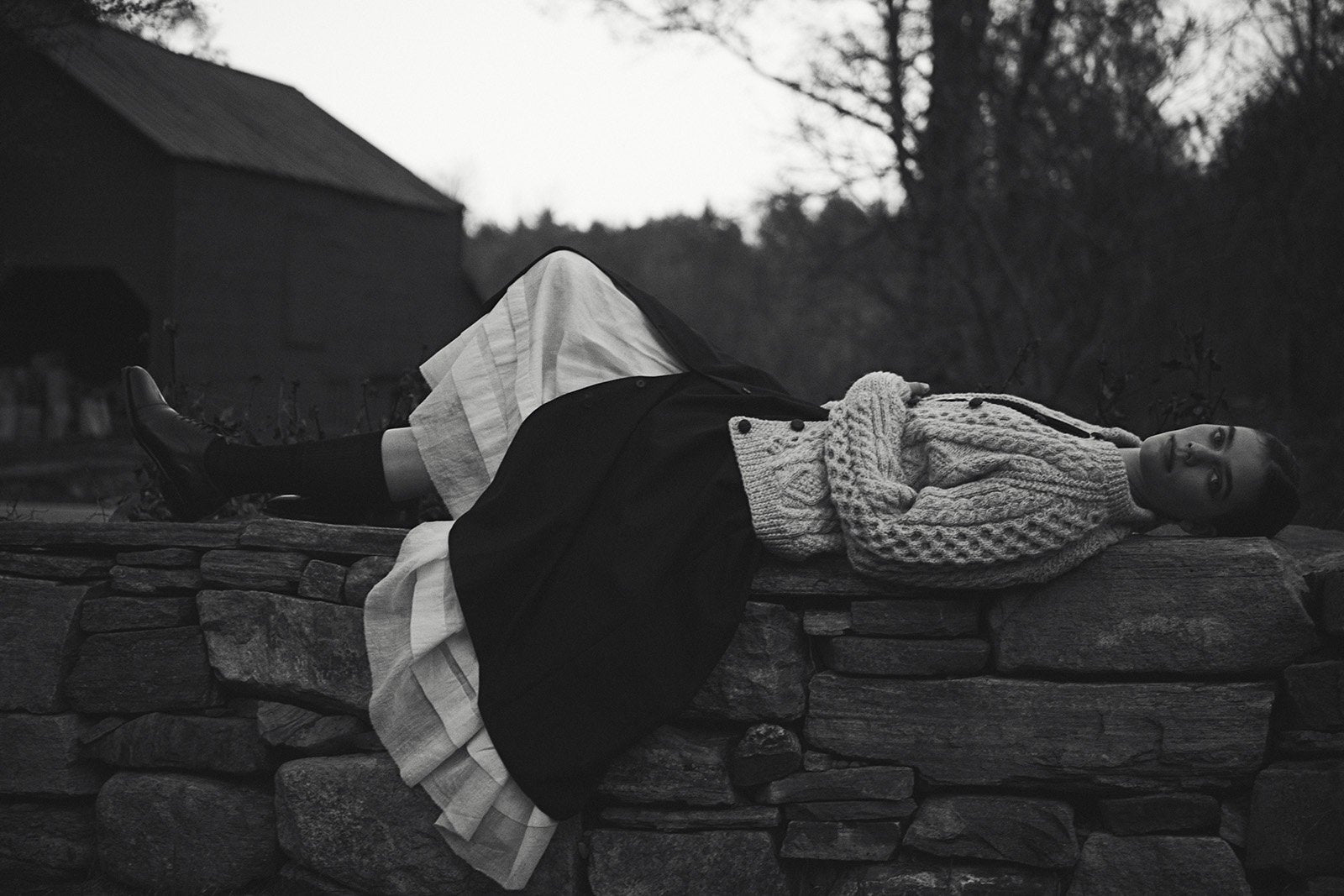 Black and white image of a model laying sideways on a stone wall wearing a long white petticoat, black skirt, and wool sweater.