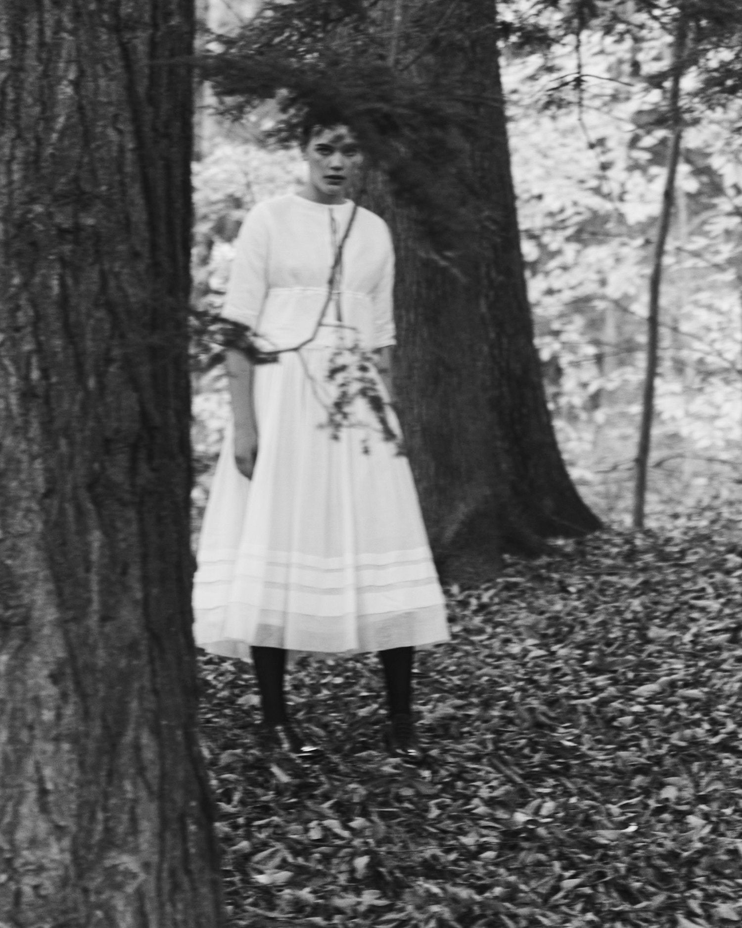 Black and white image of a girl peering out from behind a tree, wearing a long white petticoat and white top.