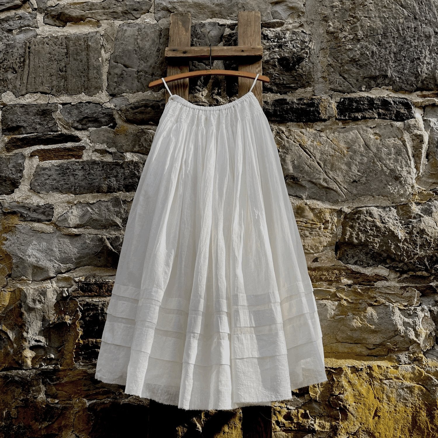 A white skirt with an elastic waitstband and french hem detailing hangs from a wooden hanger hooked to a wooden post in front of a stone wall.