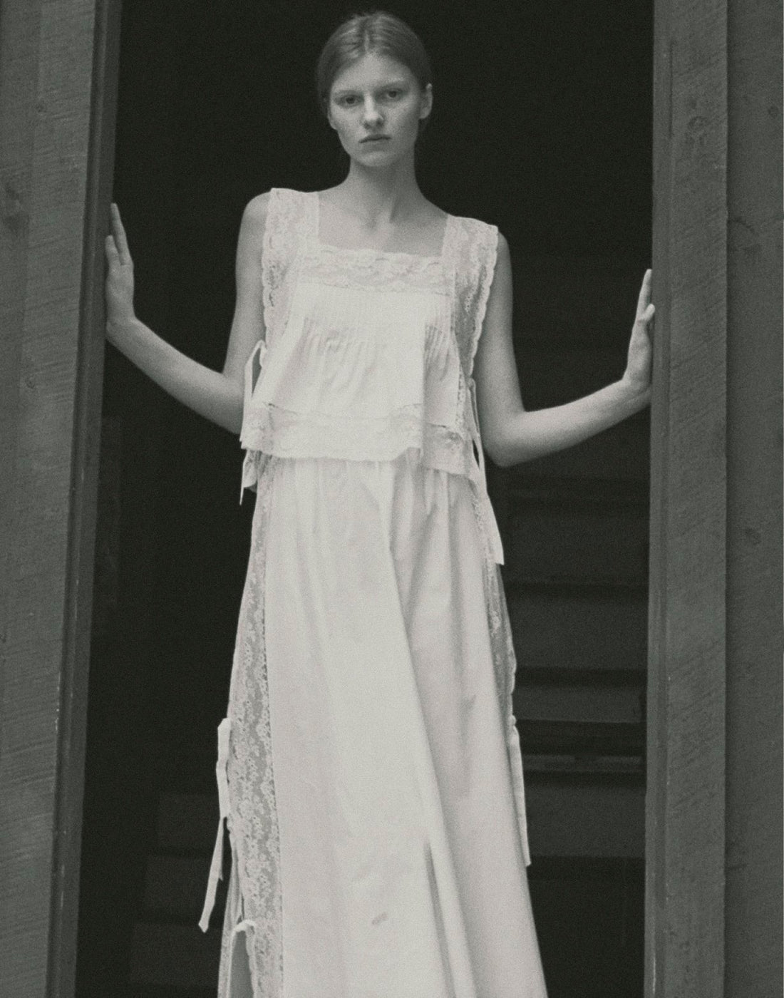 A black and white image of a model standing in a doorway wearing a white top and skirt,