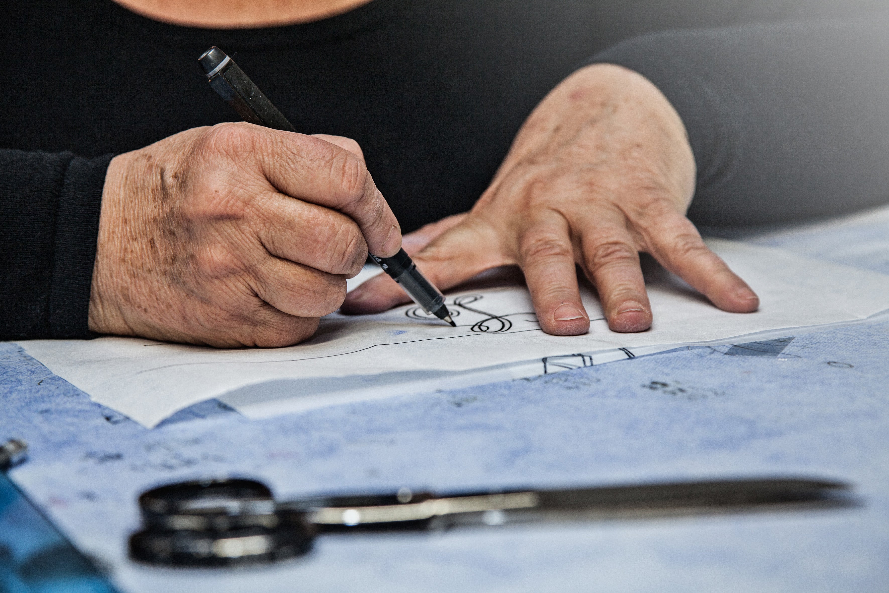 A close up image of a work desk with an artisan's hands working on monogram lettering. One hand draws a cursive L while the other hand is spread across the paper.