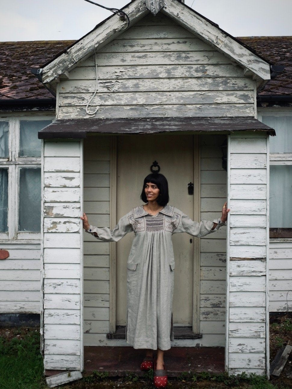 A woman standing in the doorway of an old white house wearing a dress.