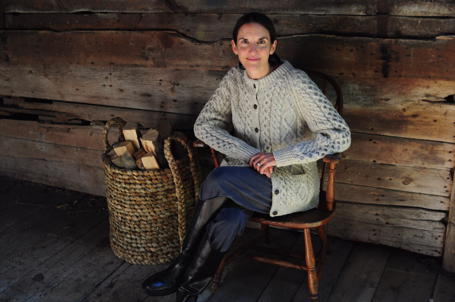 Woman seated on a porch in a grey cable knit cardign next to a woven basket of firewood.