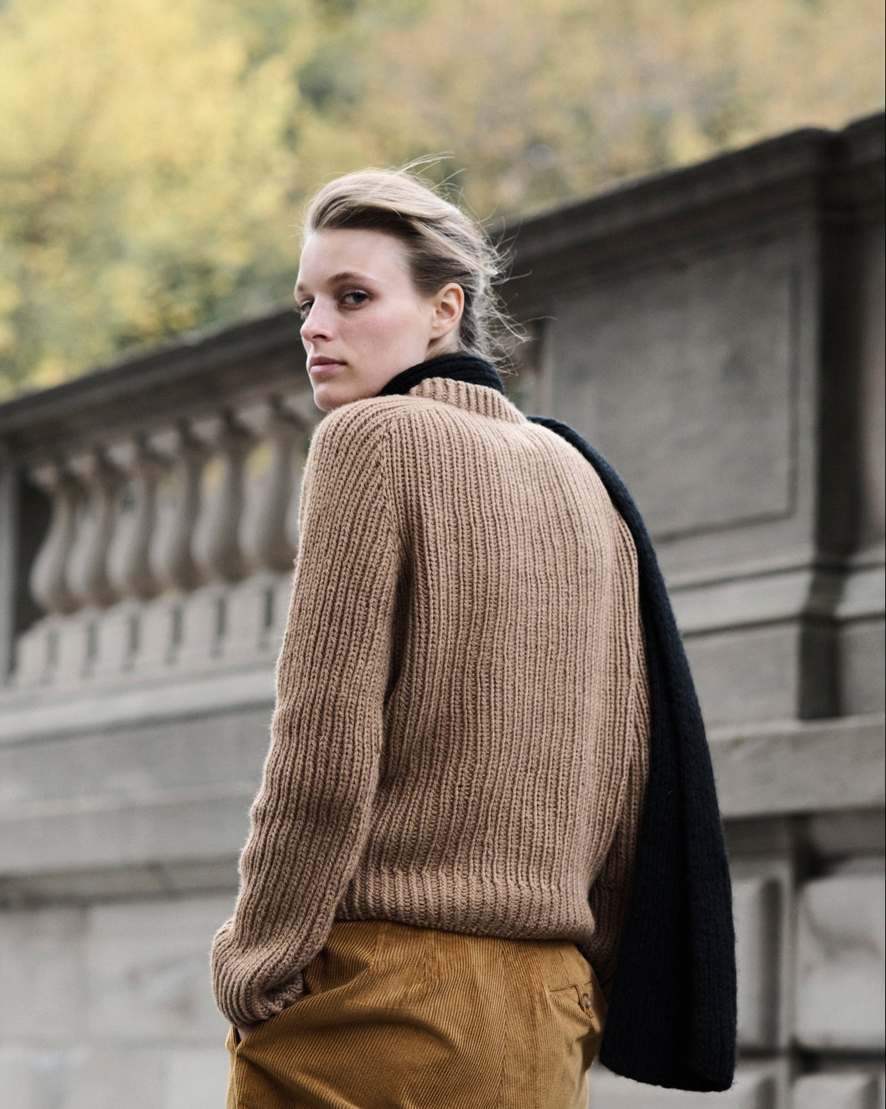 Woman wearing a beige sweater and brown pants standing on a stone bridge with trees in the background