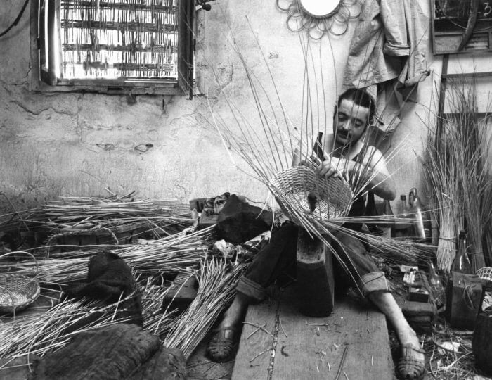 Black and white photograph of a man seated weaving a basket with rushes.