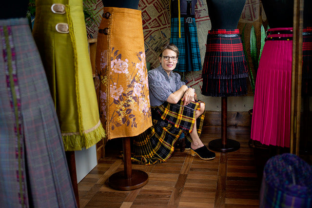 The Acme Atelier founder smiling and sitting on the floor surrounded by mannequins dressed in colorful, bespoke kilts.