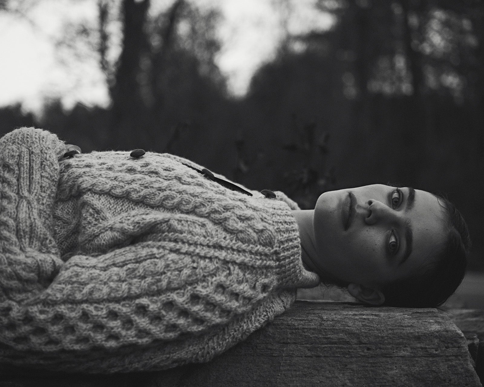 A model lays horizontally on a wooden bench. She wears a thick wool cardigan and has her head turned to look toward the camera.