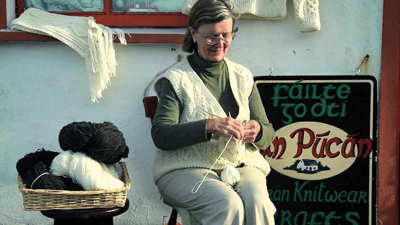 A women wearing glasses and a sweater sits in front of a white building and next to a basket stacked full of yarn while she knits.
