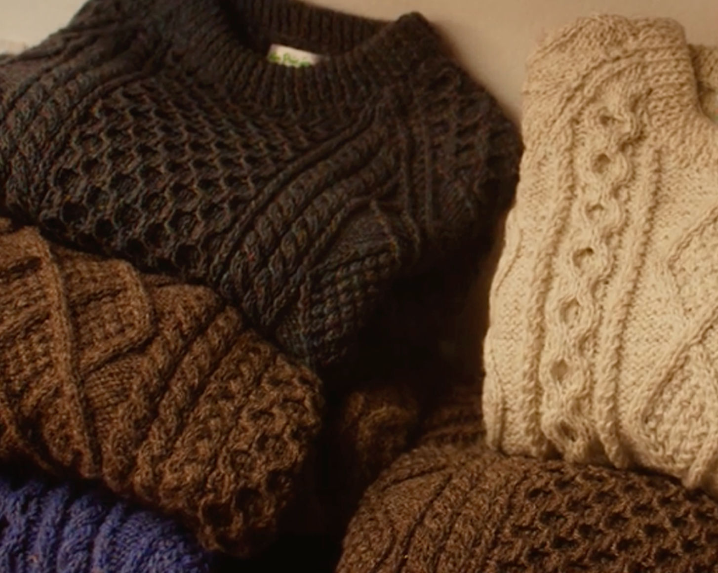 A close up photo of seven Aran cardigans in different tones of brown and cream folded on top of one another.