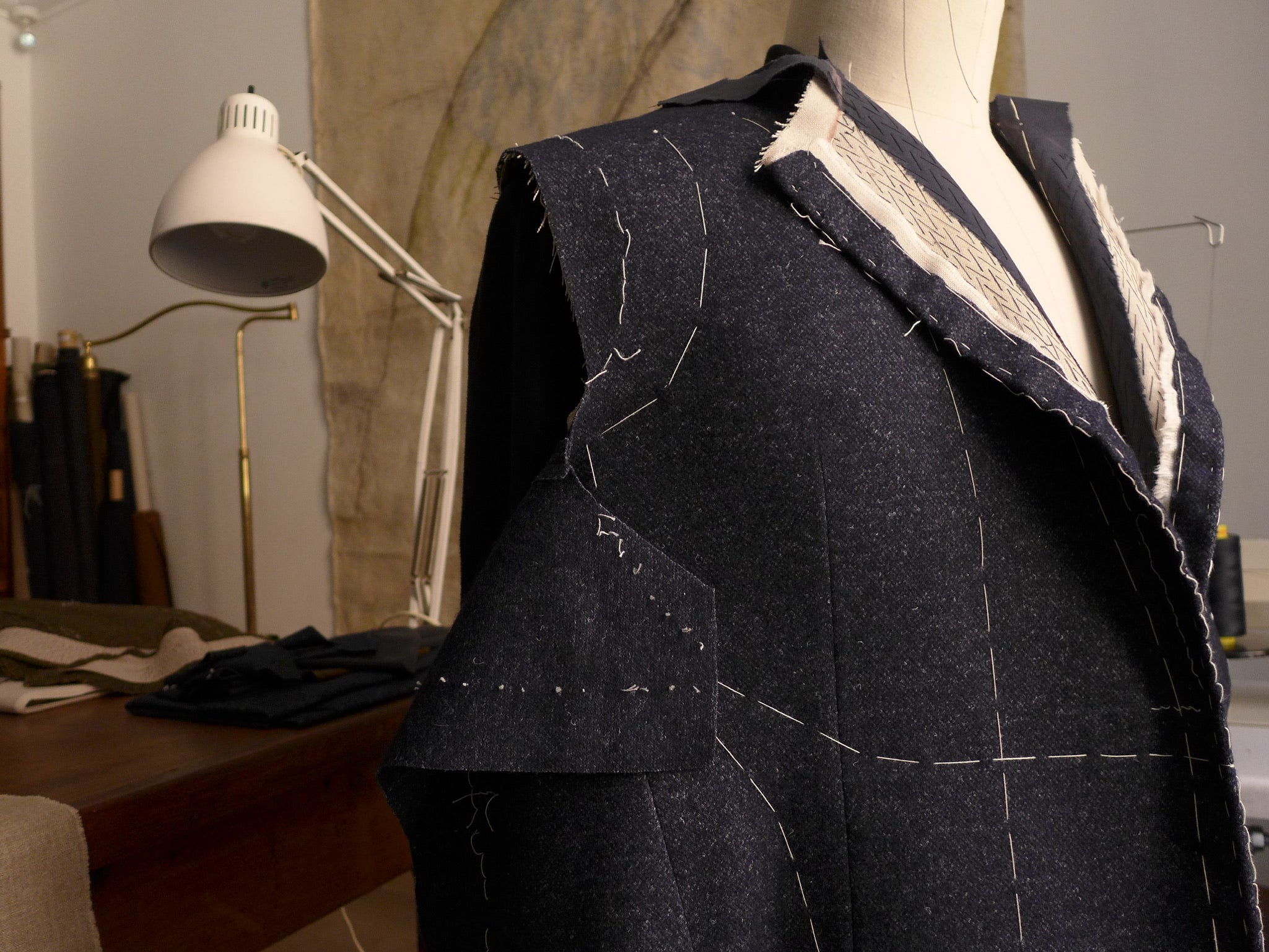 Dark grey fabric is draped on a mannequin with stitching details to guide the artisans on where to sew. The background is a studio with fabric bolts propped in a corner.