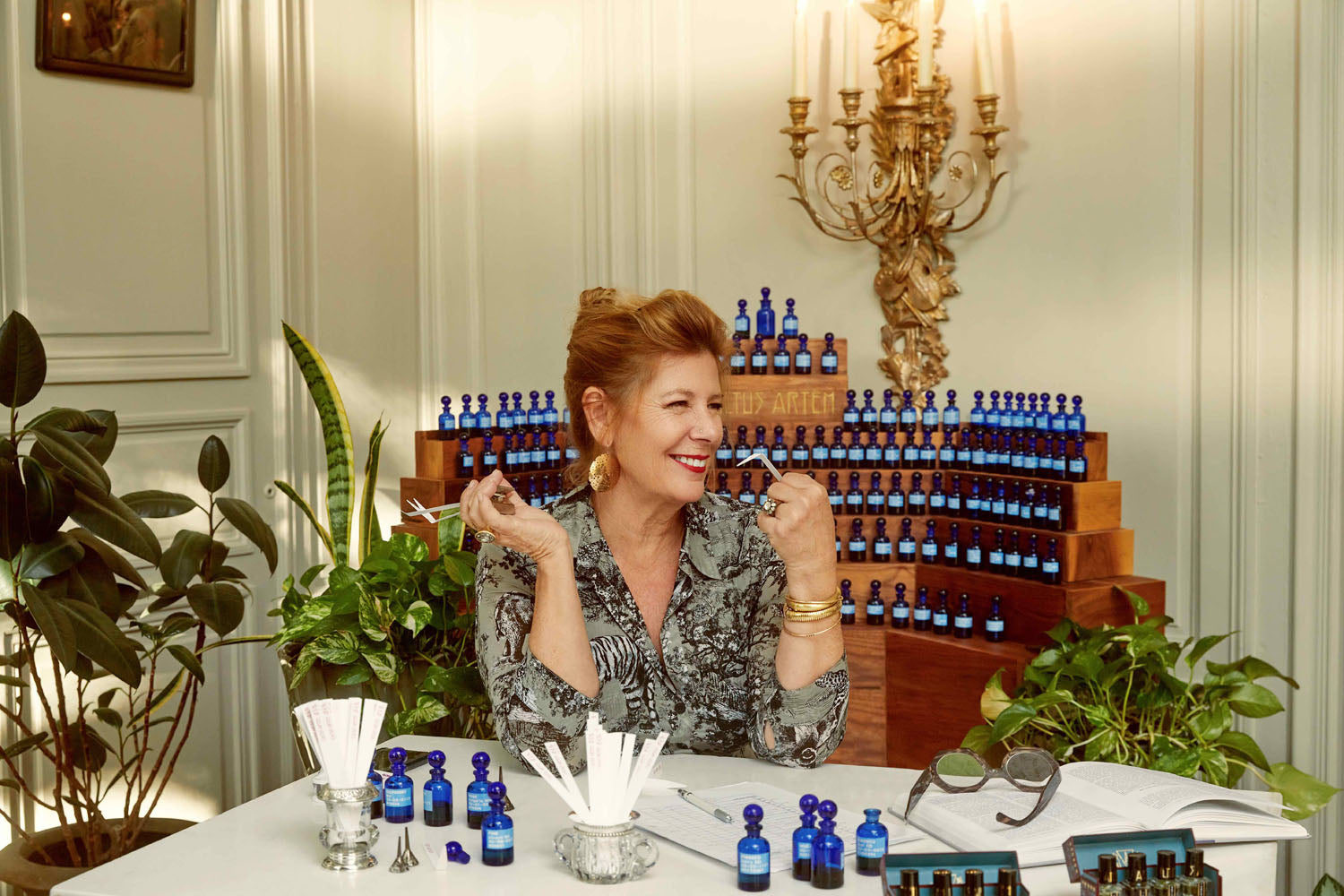 A woman with red hair and large gold earrings smiles as she sniffs scents at a table full of perfume.