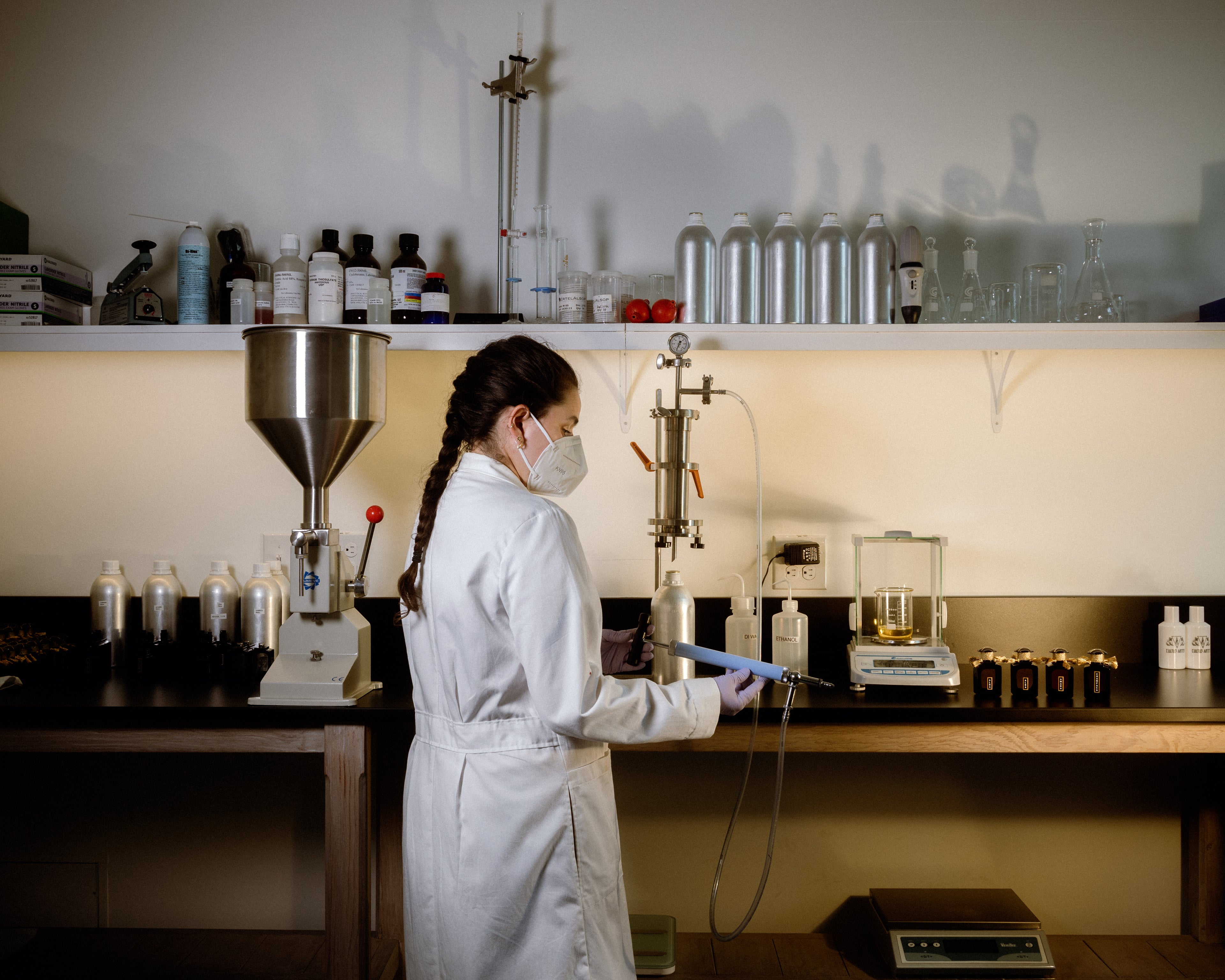 A woman with braided hair in a white lab coat and mask holds a metal tool used for making perfume in a laboratory.