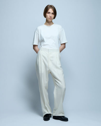 A model stands posing with her hands behind her back wearing a white t-shirt and white pleated trousers.