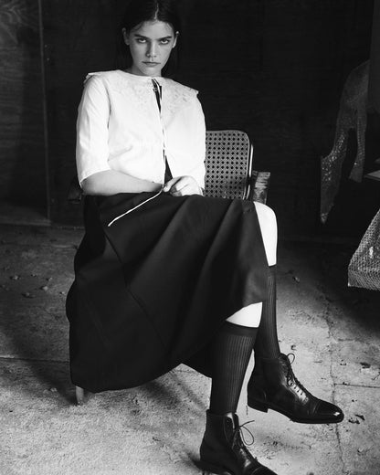 Black and white image of a model sitting cross legged, wearing a white chemise top, black skirt, and black boots.
