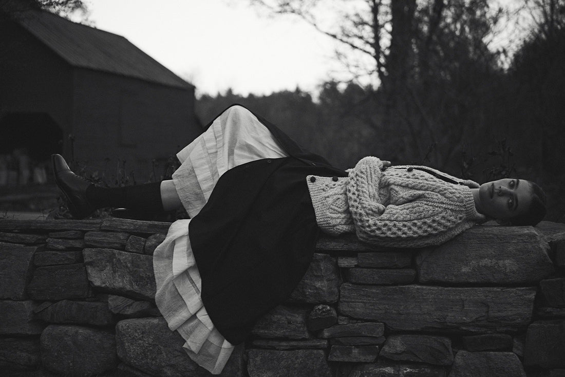 Black and white image of a model laying sideways on a stone wall wearing a long white petticoat, black skirt, and wool sweater.