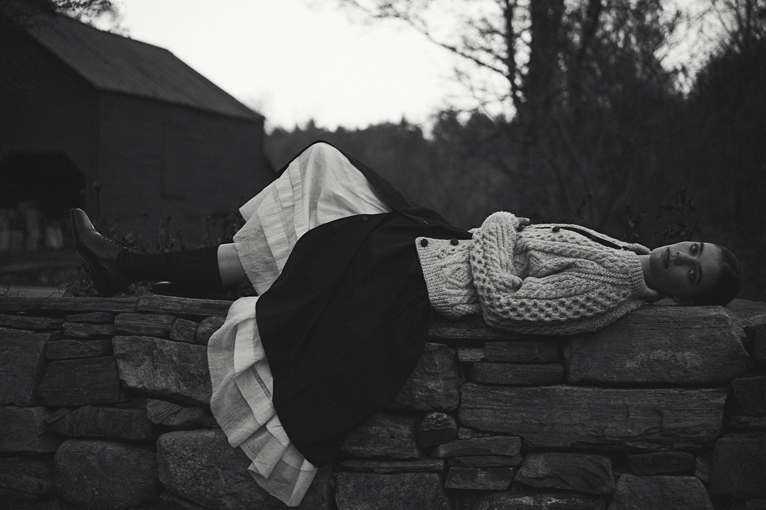 Black and white image of a model laying sideways on a stone wall wearing a long white petticoat, black skirt, and wool sweater.