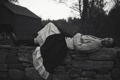 Black and white image of a model laying sideways on a stone wall wearing a long white petticoat, black skirt, and wool sweater.
