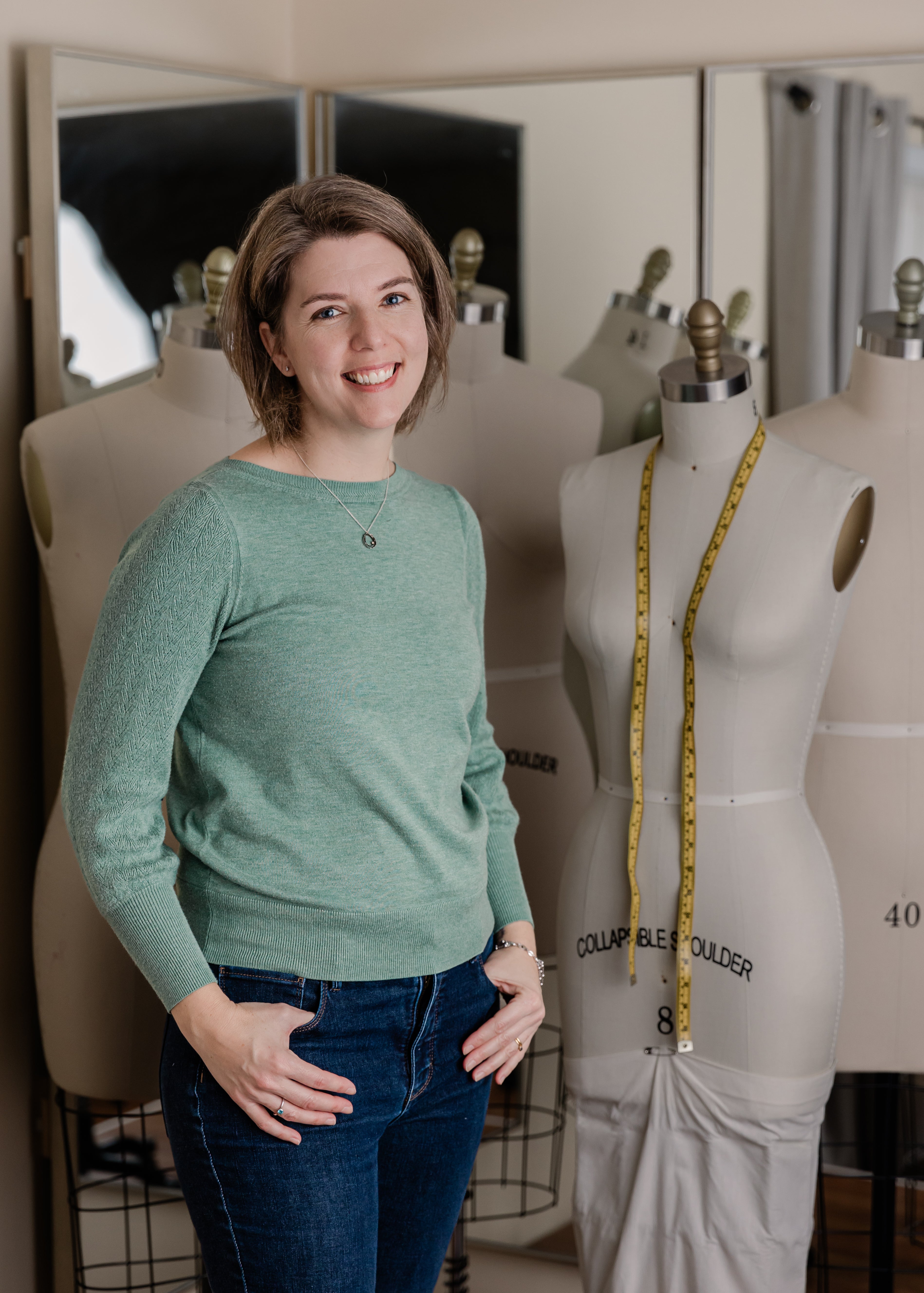 A woman in a green shirt stands smiling with her hands in her denim pant pockets next to a mannequin that has a measuring tape draped around its neck.