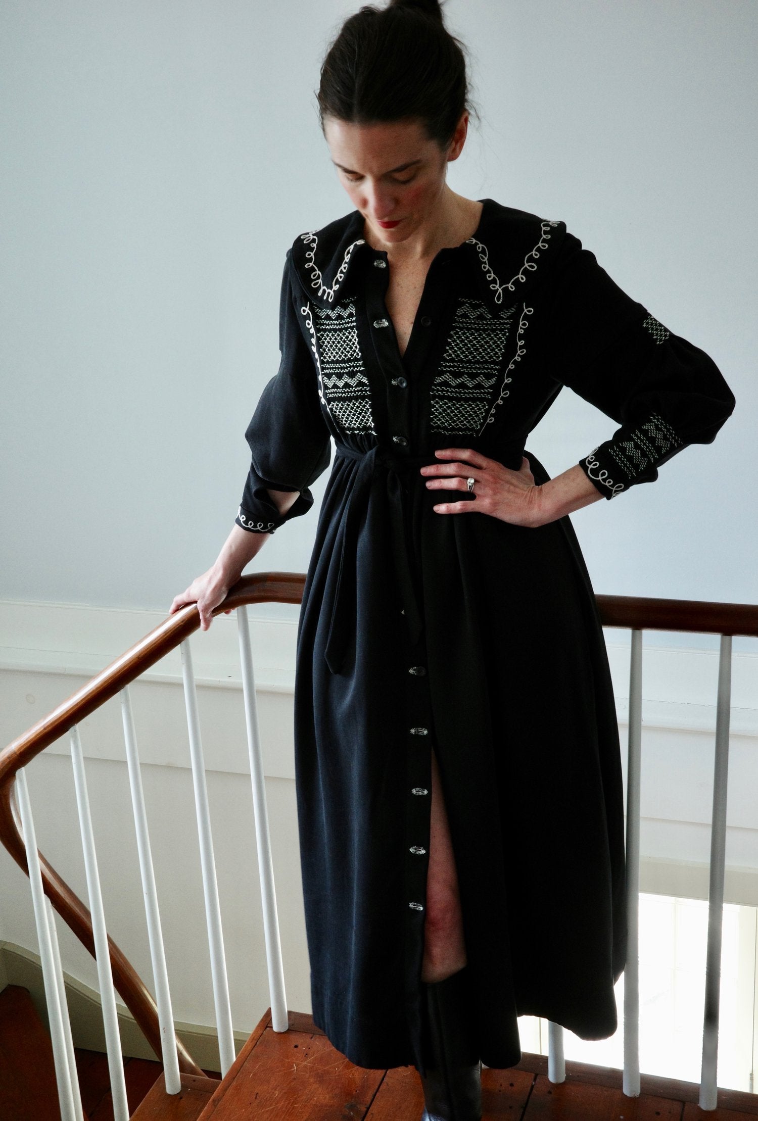 A woman standing in a stairwell wearing a black dress with white embroidery.