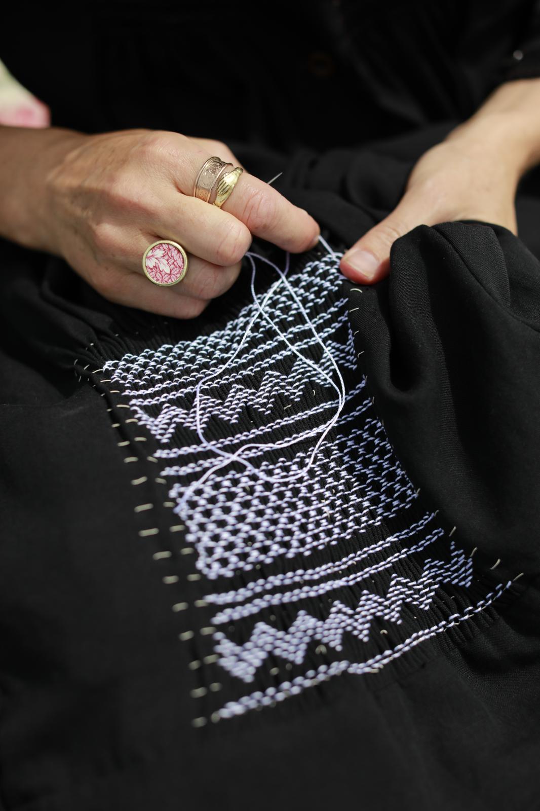 An action shot of hands adorned with rings sewing white smocking detail onto a black garment.