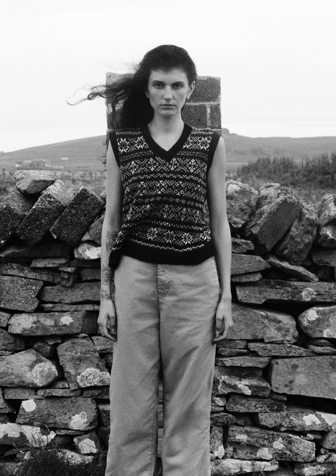 A person wearing a traditional Fair Isle patterned vest standing in front of a stone wall in a rural setting.
