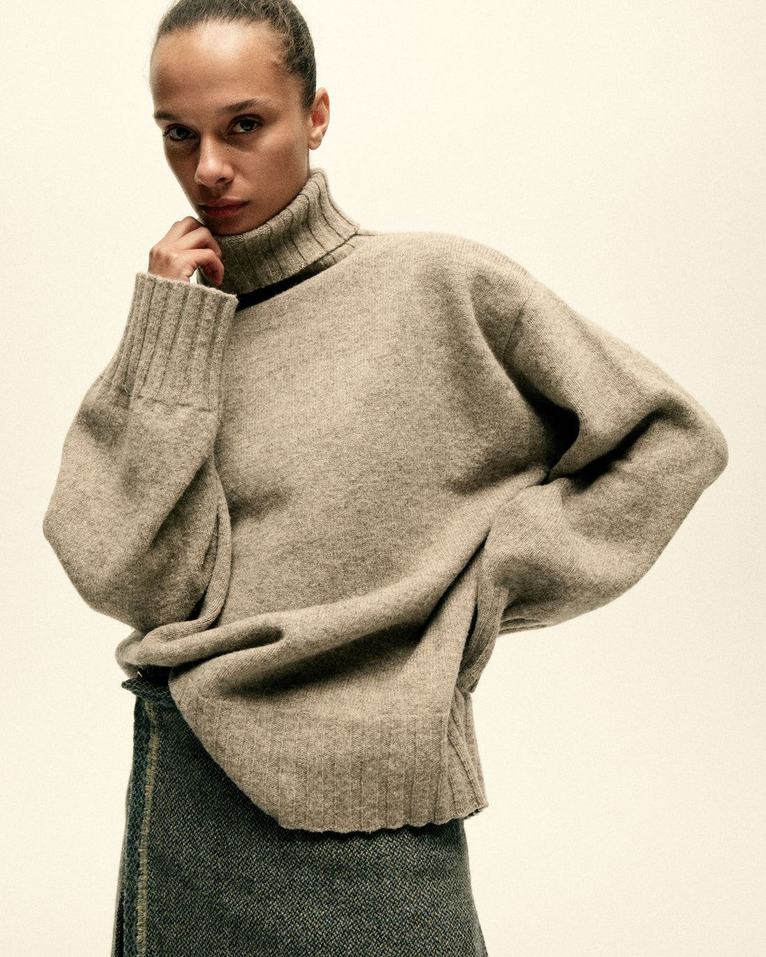 A model stands with one hand on her hip and her other hand holding her brown turtle neck sweater.