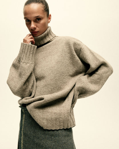 A model stands with one hand on her hip and her other hand holding her brown turtle neck sweater.