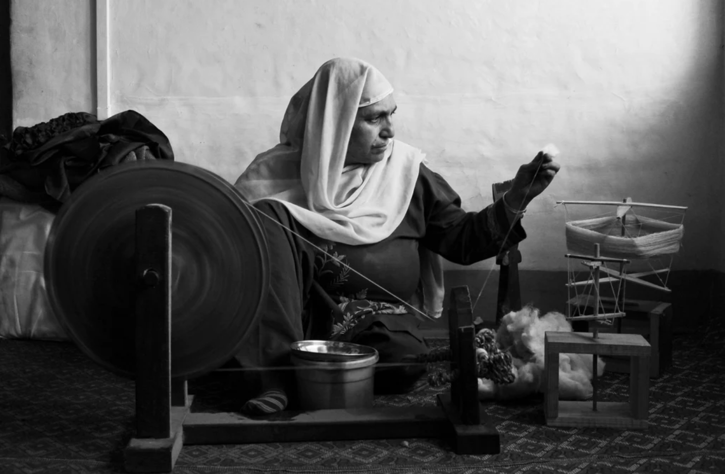 A black and white image of a woman holding holding a long thread up at eye level as she sets up a traditional loom.