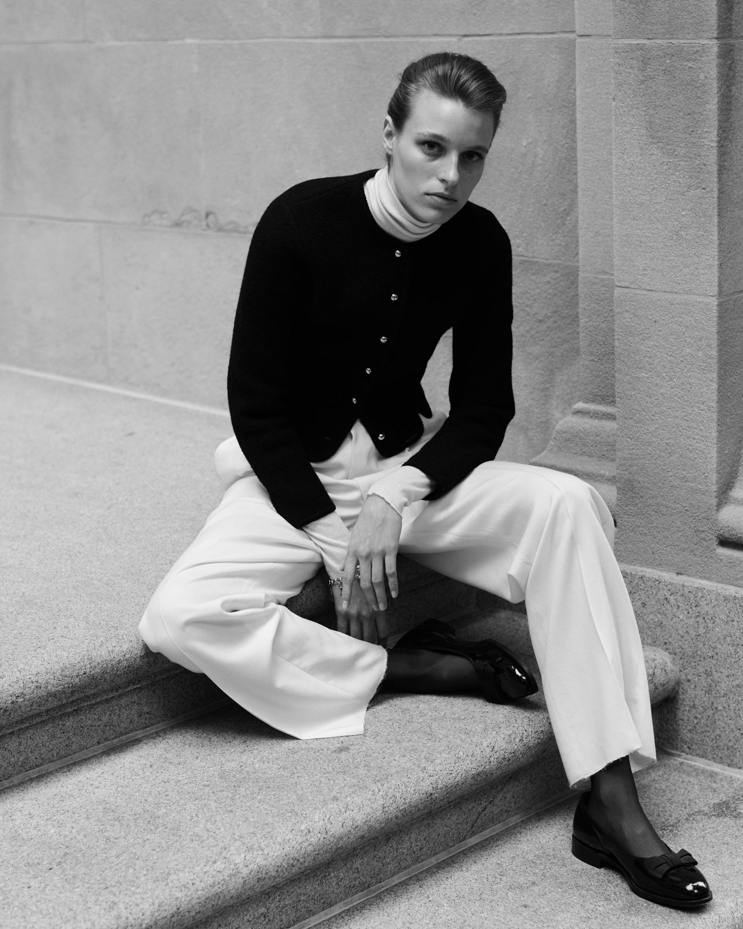 Person sitting on steps wearing a black cardigan and white pants in a black and white photo.
