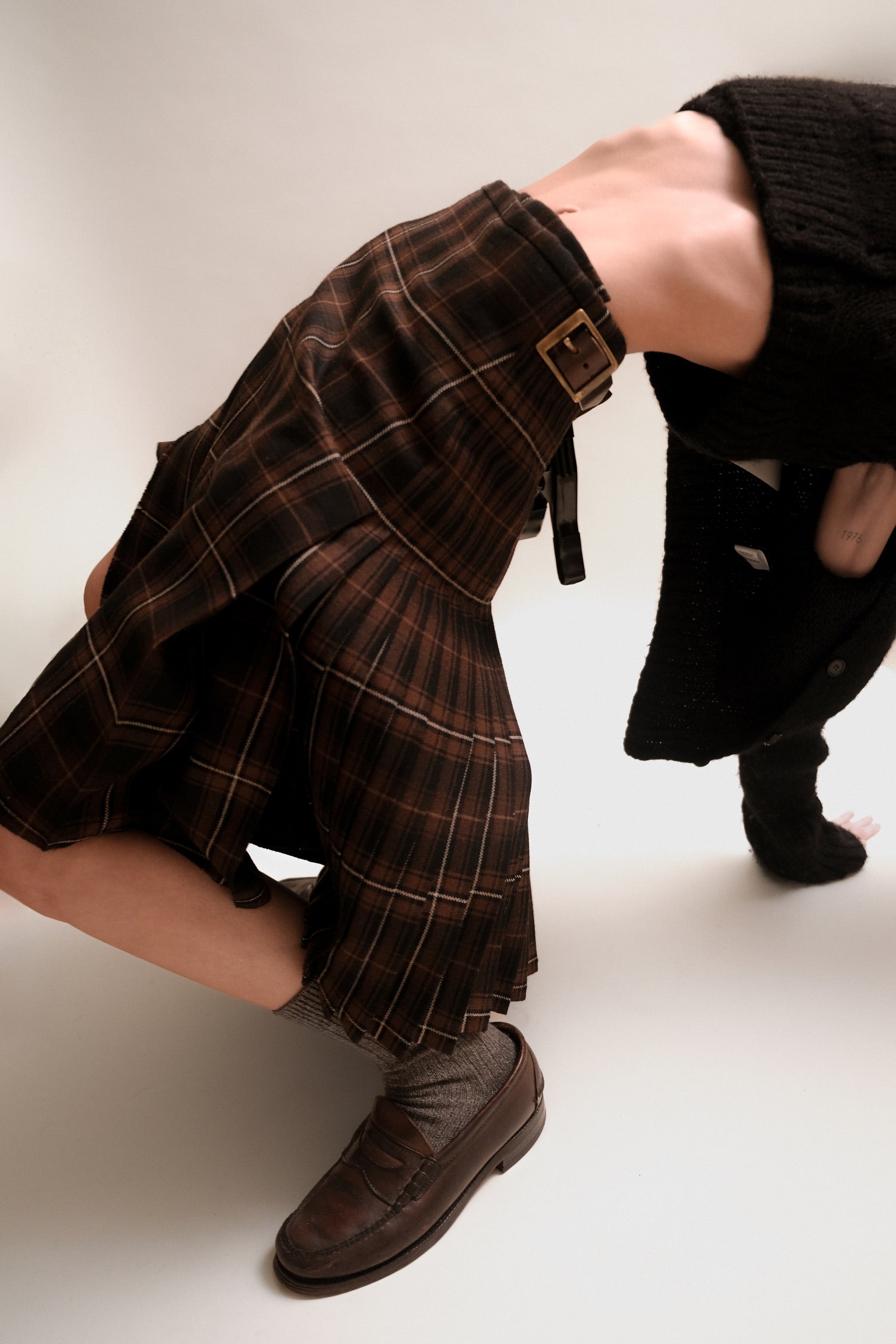 A model does a bridge pose wearing a black and brown kilt, grey socks, and leather loafers.