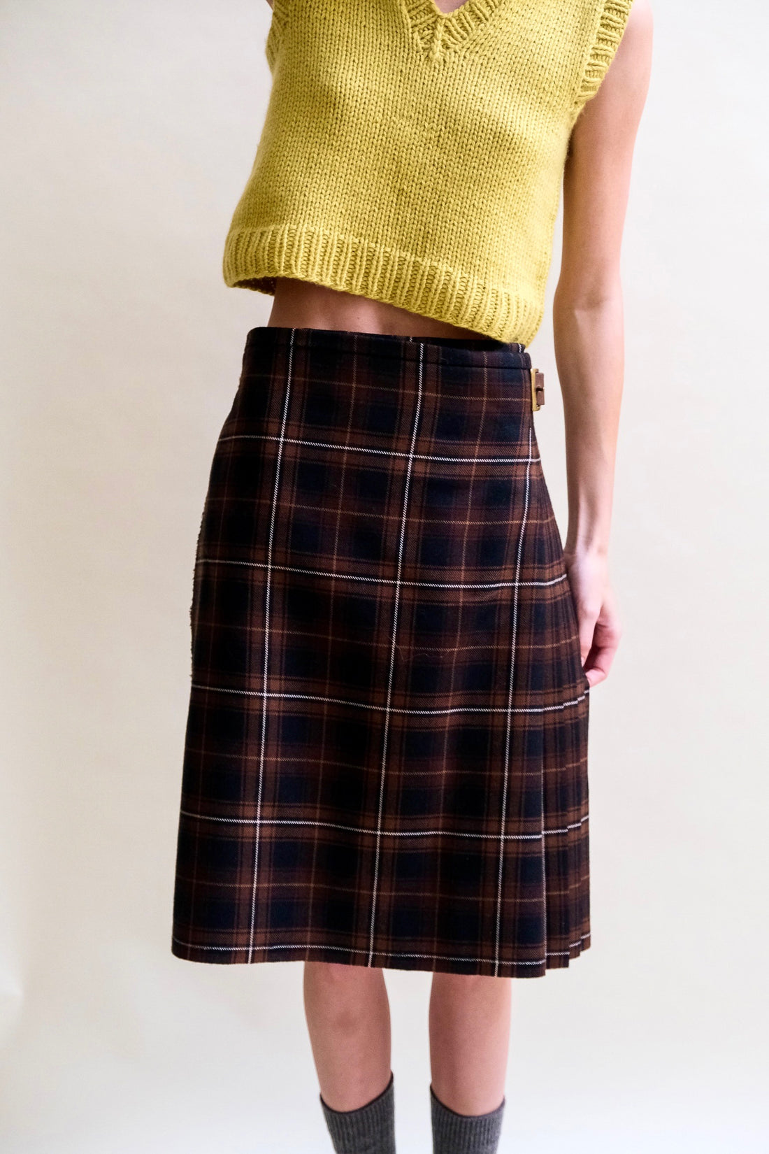 A model stands wearing a black and brown tartan kilt, chartreuse sweater vest, and grey socks.