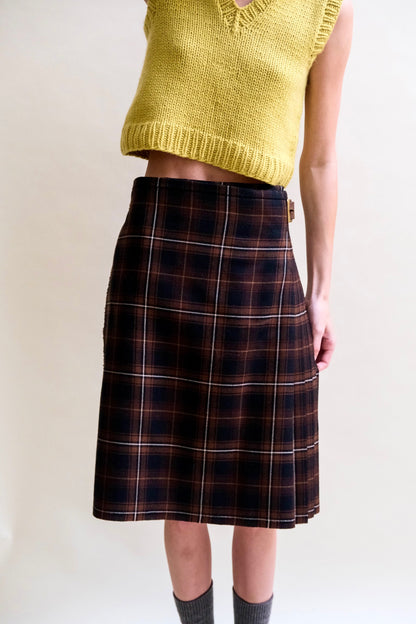 A model stands wearing a black and brown tartan kilt, chartreuse sweater vest, and grey socks.
